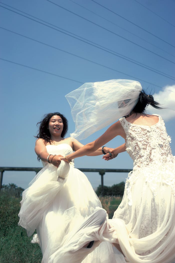 Two brides joyfully dancing in white dresses under a clear sky, showcasing happiness and celebration.